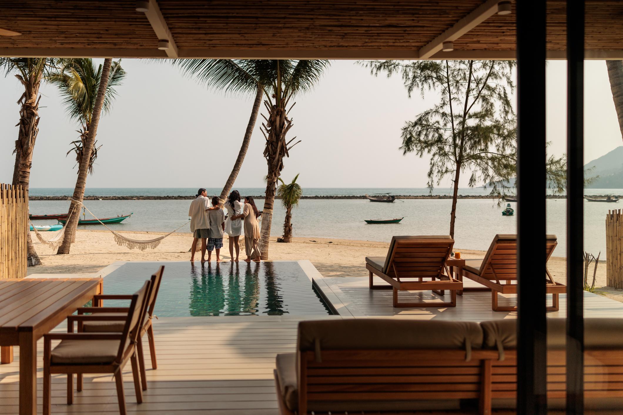 A joyful family shares a warm embrace beside the pool in their villa’s, gazing at the beautiful view of the beach and the sparkling sea in the background, in Koh Phangan, in Thailand.
